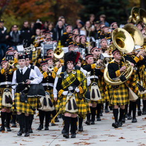 ϲʿֱֳ marching band marches in uniform with alumni and families trailing behind.